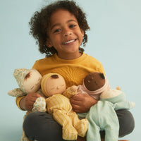 A smiling young child with curly hair sits against a light blue background, holding three soft plush dolls dressed in pastel clothing.
