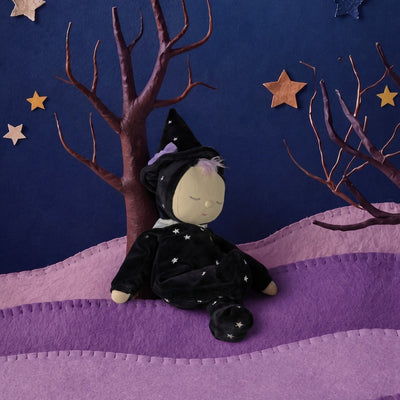 A plush witch doll wearing a black pointed hat and starry dress sits on purple fabric hills under a dark blue backdrop with paper stars and bare tree branches.
