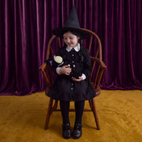 A young girl wearing a black witch costume with a pointed hat, white collar, and black dress sits on a wooden chair in front of a purple velvet curtain and mustard-yellow floor, holding a small black doll.