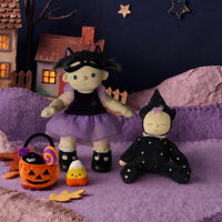 A Halloween-themed miniature scene with two fabric dolls dressed as witches, an orange jack-o'-lantern bucket, a candy corn, and autumn leaves on a purple surface against a dark blue night sky backdrop with stars and a crescent moon.