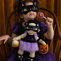 A young girl in a purple and black cat-themed costume sits in a wooden chair holding a matching plush doll, with a pumpkin bucket and a lollipop against a golden-brown curtain backdrop.