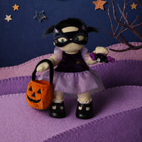 A plush doll dressed as a black cat in a purple costume stands on a purple felt landscape, holding an orange pumpkin bucket and a purple toy. The background features a dark blue felt sky with a yellow felt crescent moon, star cutouts, and a bare tree branch.