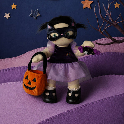 A plush doll dressed as a black cat in a purple costume stands on a purple felt landscape, holding an orange pumpkin bucket and a purple toy. The background features a dark blue felt sky with a yellow felt crescent moon, star cutouts, and a bare tree branch.