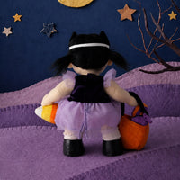 A plush doll viewed from behind wearing a black and lavender Halloween costume, holding an orange pumpkin and a purple bucket, standing on a purple hill against a dark blue night sky backdrop with stars and a crescent moon.