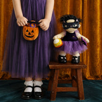 A child wearing a purple tulle Halloween costume skirt and black top stands beside a small wooden stool holding a plush doll dressed in a black mask and purple outfit, with an orange pumpkin bucket bag. The background is a gold curtain and dark teal floor.