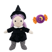 A plush witch doll with a black hat and dress, purple hair, and striped black-and-white socks, shown next to a small purple candy-shaped plush toy with orange ends and a yellow crescent moon on a plain white background.