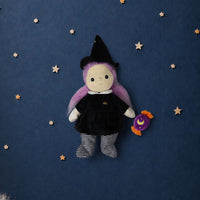 A plush witch doll with purple hair, a black dress, and striped socks sits against a dark blue starry background with white clouds at the bottom.