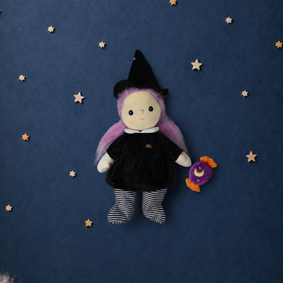 A plush witch doll with purple hair, a black dress, and striped socks sits against a dark blue starry background with white clouds at the bottom.