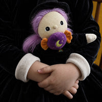 A child wearing a black velvet witch costume with a white Peter Pan collar holds a small plush toy with purple hair, a black hat, and a purple moon-shaped pillow.