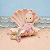 A mermaid doll with a pink dress and iridescent tail sits inside a large pink clam shell on a sandy beach set with blue ocean waves and pearls.