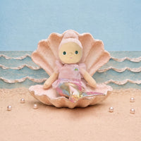A doll with a pink hat and pink dress sits inside a large pink clam shell on a sandy beach set with a blue textured background and small pearls.