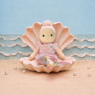 A doll with a pink hat and pink dress sits inside a large pink clam shell on a sandy beach set with a blue textured background and small pearls.