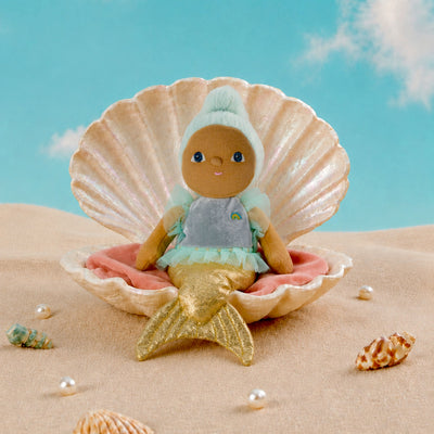 A small mermaid doll with brown skin, blue eyes, a mint green hood, and a gold tail sits inside a large beige seashell on pink sand, with a blue sky backdrop and scattered shells around.