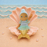 A mermaid doll with a blue and gold tail sits inside a large pink clam shell on a sandy beach backdrop with blue ocean waves behind it.