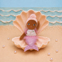 A small mermaid doll with brown skin and blue eyes sits inside a large beige seashell on a sandy surface, with a blue ocean backdrop and scattered pearl-like beads.