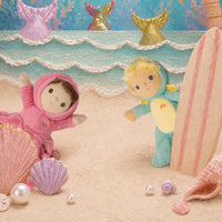 Two small plush mermaid dolls on a sandy beach scene with shells, pearls, a surfboard, and mermaid tail decorations in the background.