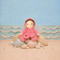 A plush doll with brown hair sits on rocks in a beach-themed diorama, wearing a pink hooded outfit against a blue ocean and sky backdrop.
