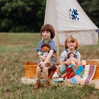 Two children with dolls sitting on a wooden toy boat in a grassy field with a teepee in the background.