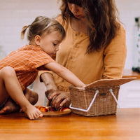 Piki Rattan Basket - Natural with white straps beside a child and adult preparing food at a table by Olli Ella Kids Rattan