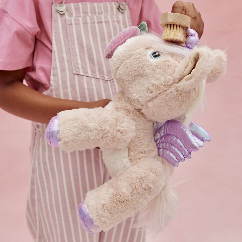 Dinkum Unicorn - Shimmer plush pink unicorn toy with purple wings and mane held by a child in a striped apron from Dinkum Pets