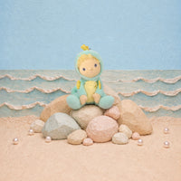A plush doll with a yellow face and blonde hair sits on a stack of rocks on a sandy beach set. The doll is wearing a light blue hooded outfit with a yellow belly and small yellow accents.