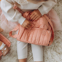 Carrie Convertible Changing Set - Rose by Dinkum Dolls shown as a soft pink doll changing bag with straps and pockets beside a soft doll on a fluffy rug