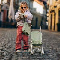 Carrie Convertible Changing Set - Sage by Dinkum Dolls with a child carrying a doll beside a sage doll pram on a cobbled street