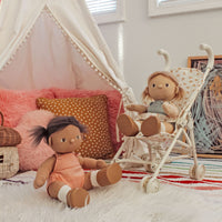 Dinkum Dolls - Gigi soft doll with golden hair in a powder-blue romper seated in a toy pram beside another doll in a cosy playroom