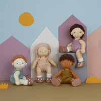 Four wooden dolls with painted faces and clothing are arranged in front of stylized pastel house shapes on a tan surface against a light blue background.