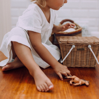 Piki Rattan Basket - Natural by Olli Ella Kids Rattan beside a child playing with a soft doll on a wooden floor