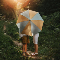 Olli Ella Kids See-Ya Umbrella - Blue Butter shelters two children on a sunlit forest path with a lightweight recycled polyester canopy for outdoor play