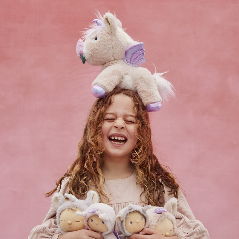 Dinkum Unicorn - Shimmer plush unicorn perched on a smiling child's head with soft dolls in her arms from Dinkum Pets