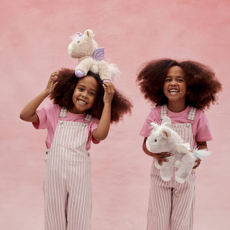 Dinkum Unicorn - Shimmer plush unicorn toy with soft wings held and balanced by smiling children in pink overalls against a pink background from Dinkum Pets