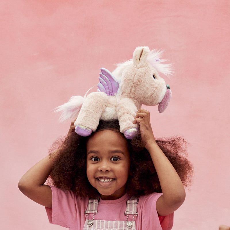 Dinkum Unicorn - Shimmer plush unicorn held above a smiling child with pink wings and soft cream fur from Dinkum Pets