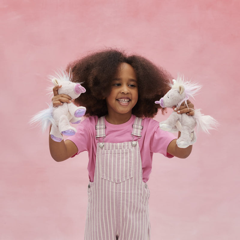 A smiling child holds two soft unicorn toys with pastel fur and glittering horns Binky Baby Unicorn - Glitter by Dinkum Pets
