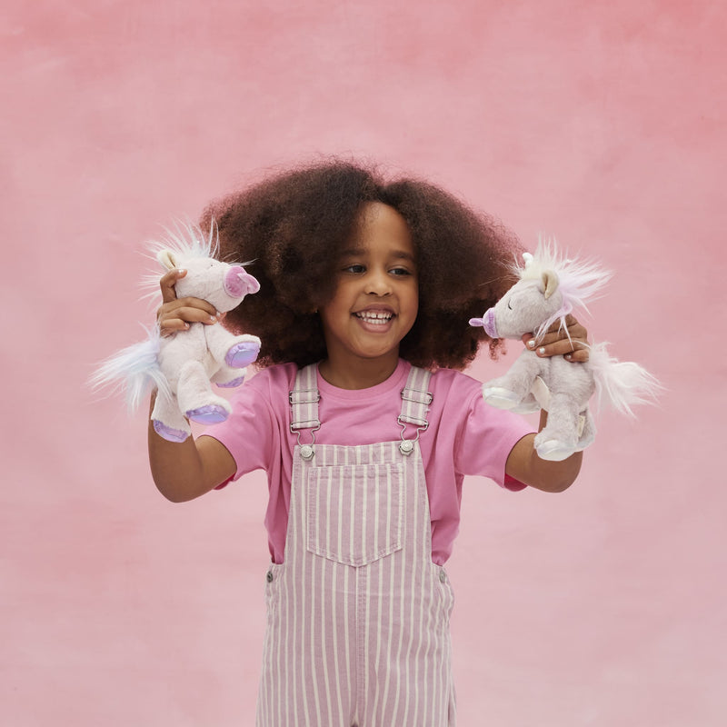 Binky Baby Unicorn - Sparkle by Dinkum Pets held by a smiling child alongside another unicorn plush toy in a playful pink setting